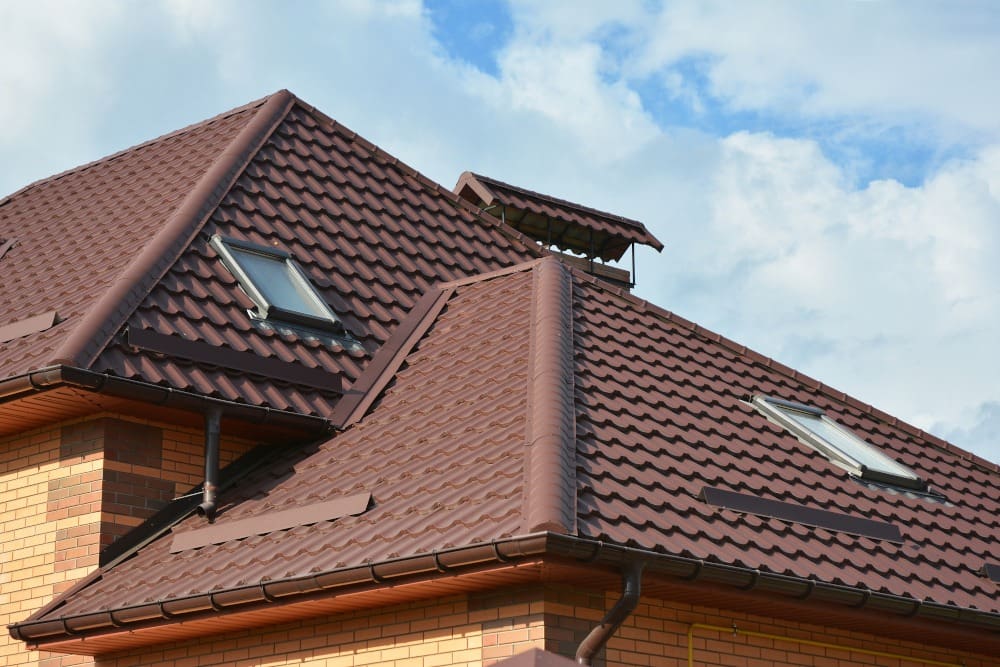 A house with a brown, tiled roof featuring multiple peaks and skylights against a cloudy sky.