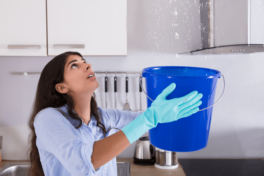 Woman in a kitchen holds a blue bucket under a leak from the ceiling, wearing blue gloves.