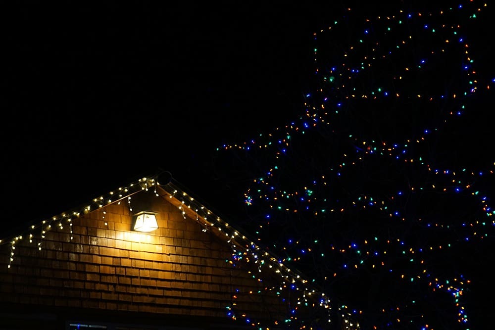 A house roof with string lights under a lit porch area. A tree nearby is adorned with colorful string lights.