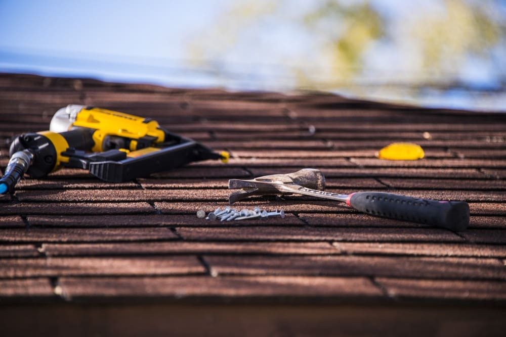 A roof with a nail gun, hammer, and nails scattered on brown shingles.