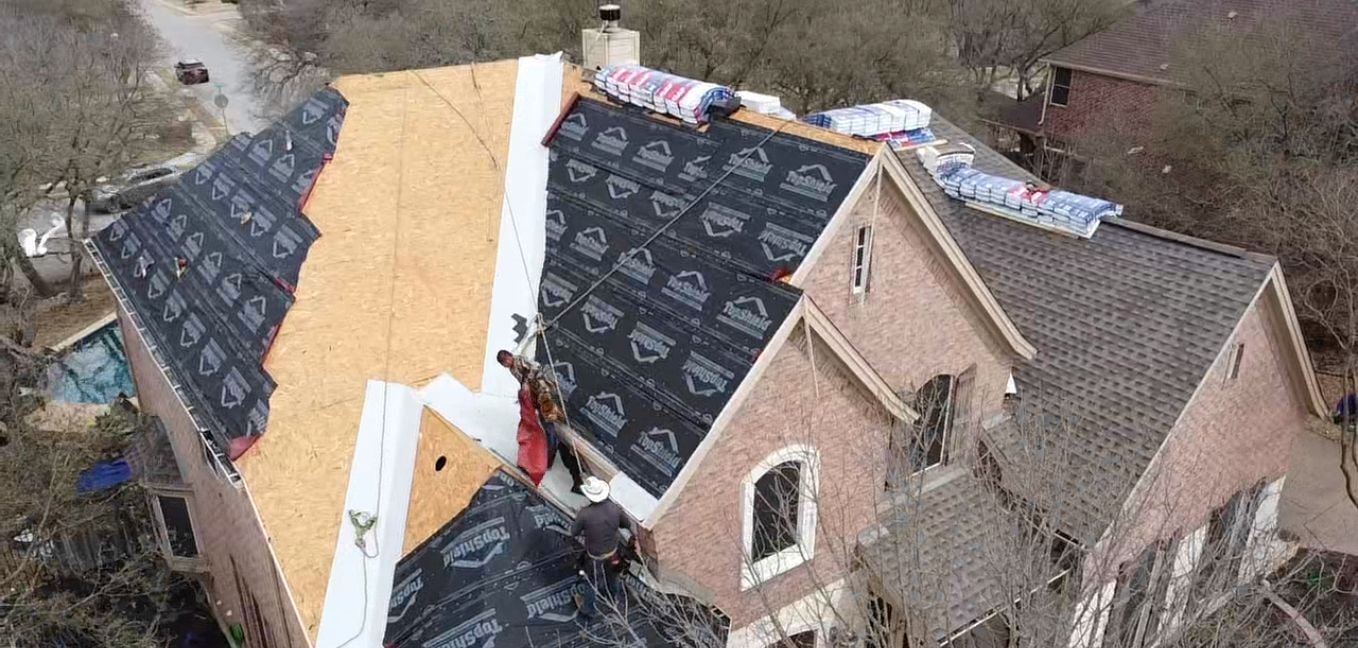 Two workers are installing roofing underlayment on a house roof, with bundles of shingles placed on the roof.