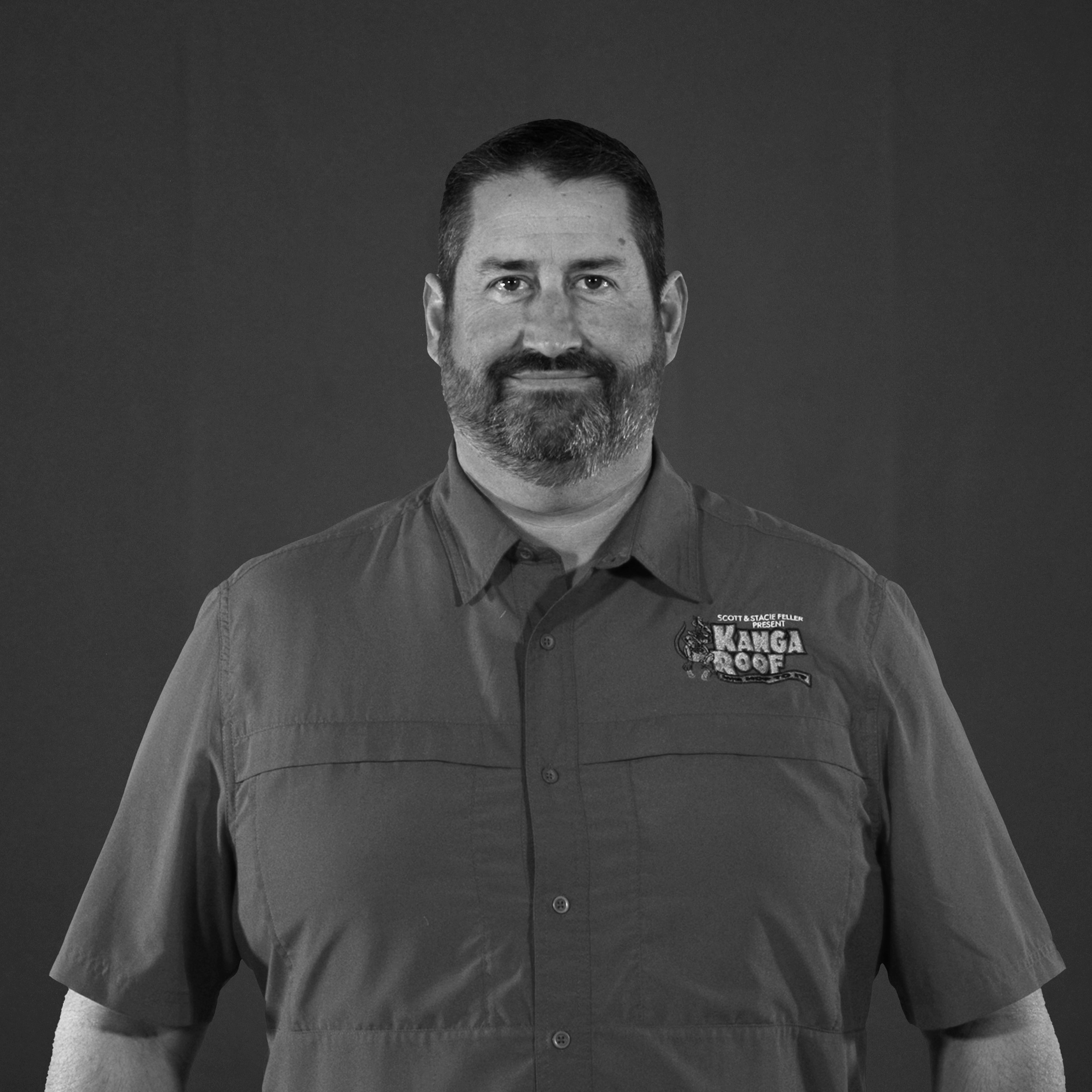 A man with a beard wearing a short-sleeve button-up shirt with a Kanga Roof logo stands against a plain dark background, facing the camera.
