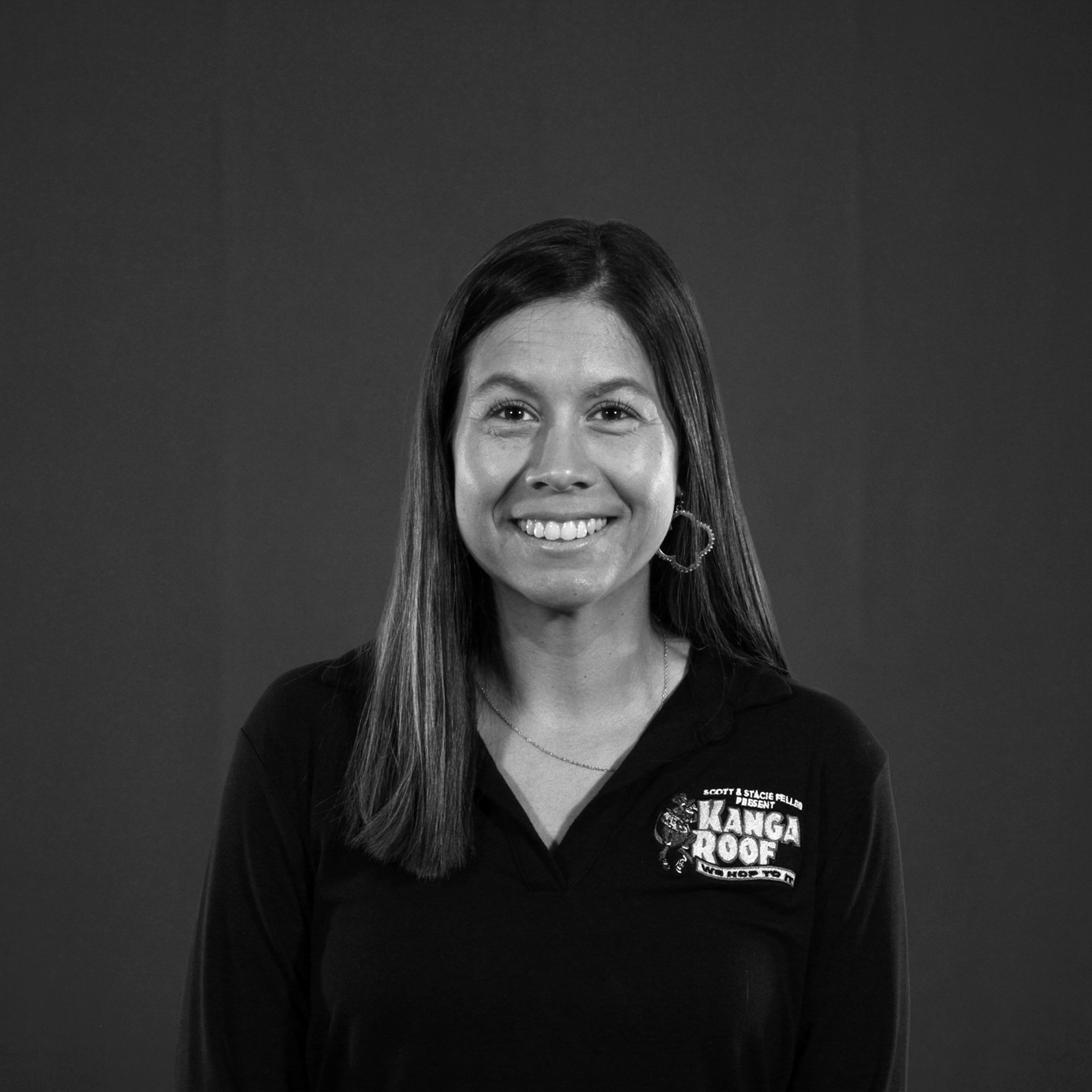 A woman with straight hair smiles at the camera, wearing a dark collared shirt with a "Kanga Roof" logo, against a plain background.