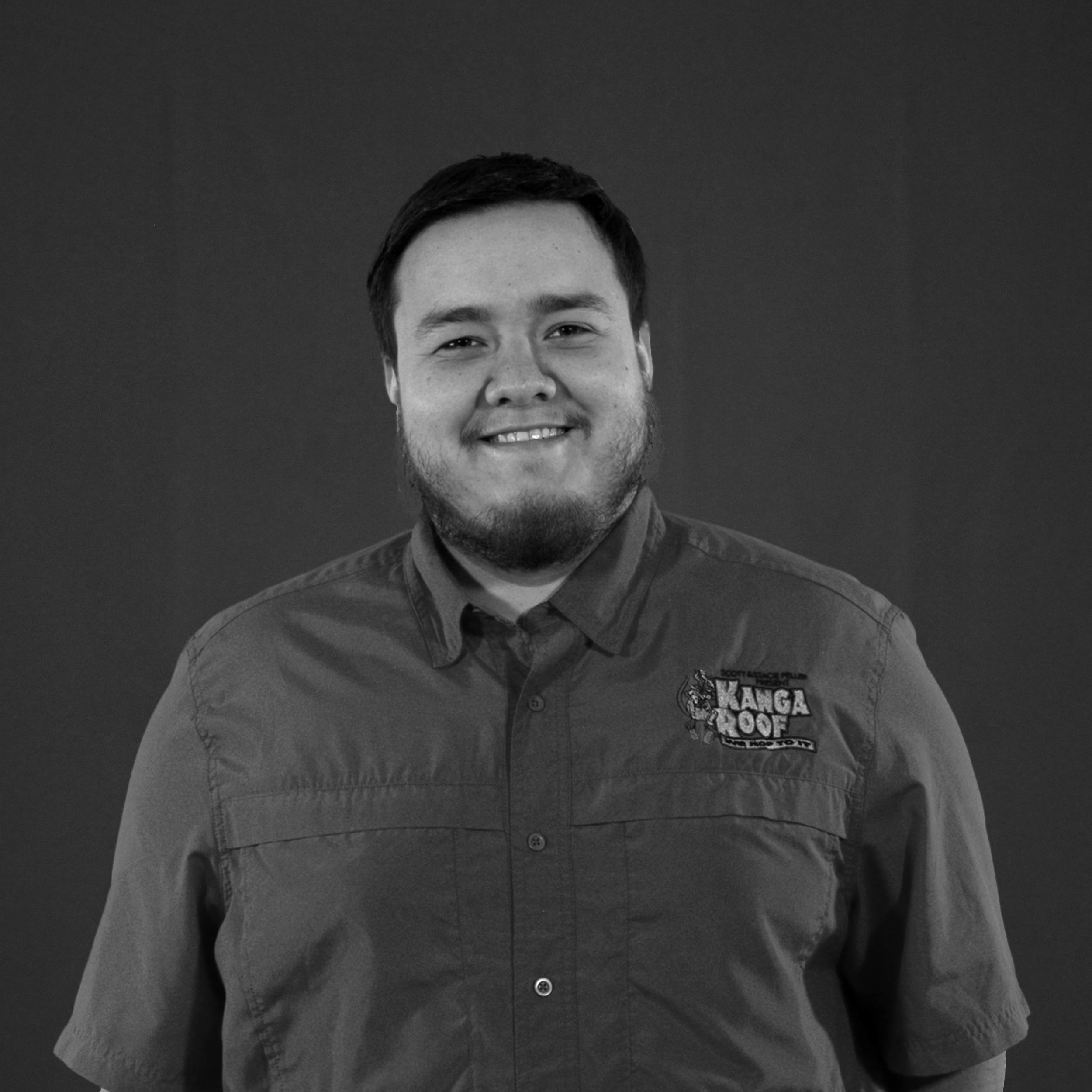 A man with short hair and a beard, wearing a "Kanga Roof" shirt, stands smiling in front of a plain dark background.