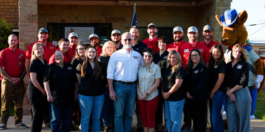 A group of kangaroof employees poses for a photo outside a building, with an American flag and a person in a kangaroo costume in the background.