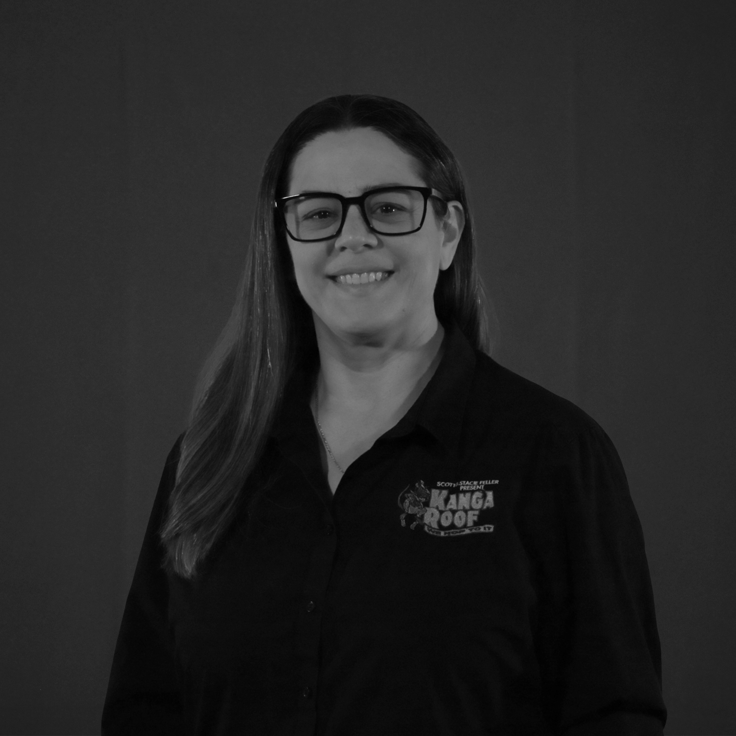 A woman with long hair and glasses smiles at the camera, wearing a dark shirt with a "Kanga Roof" logo, against a plain dark background.