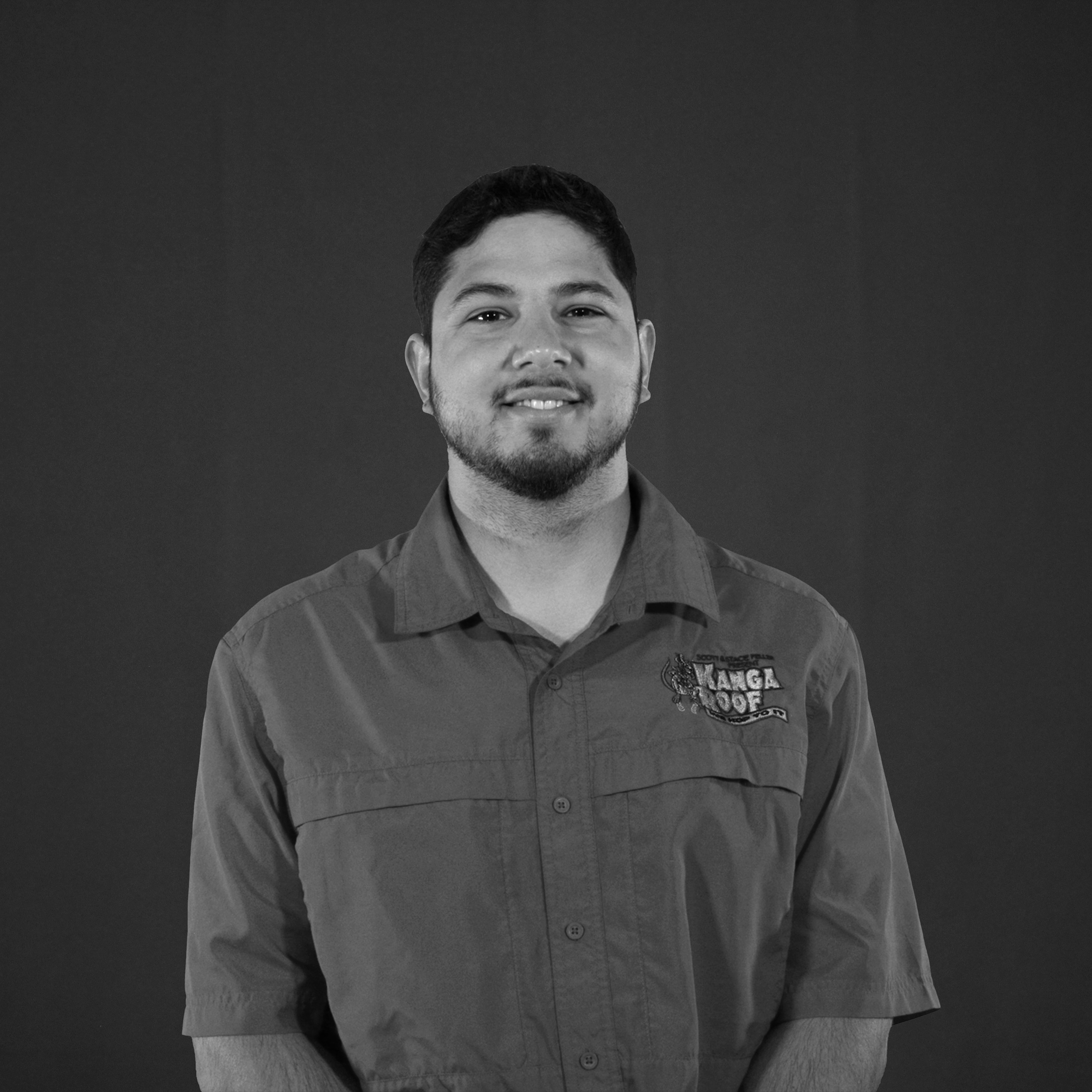 A man with short dark hair and a beard stands against a plain background, wearing a button-up shirt with a "Kanga Roof" logo.