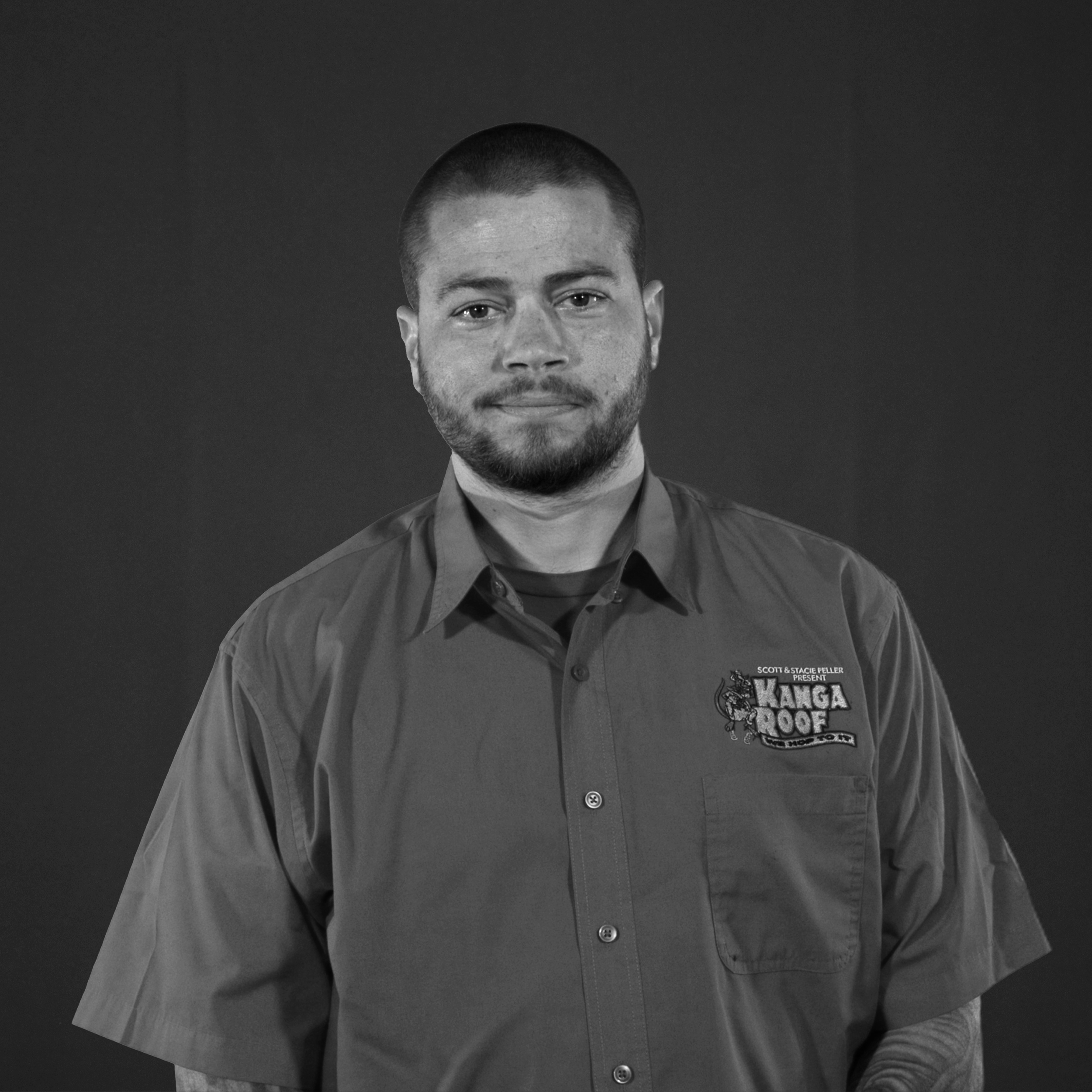 A man with short hair and a beard wears a button-up shirt with a "Kanga Roof" logo, standing against a plain dark background.