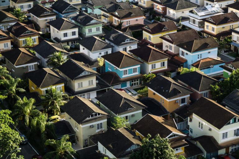 Aerial view of a densely packed neighborhood with colorful houses and dark roofs, surrounded by lush green trees.